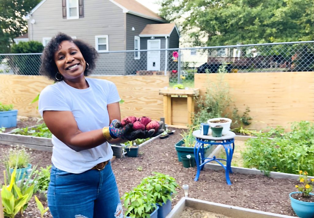 Cheryl wearing white t-shirt and jeans, holding homegrown red potatoes in her Backyard Oasis.